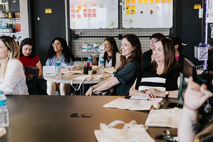 Em volta de uma mesa, algumas mulheres prestam atenção em algo que está sendo apresentado em sala de aula.