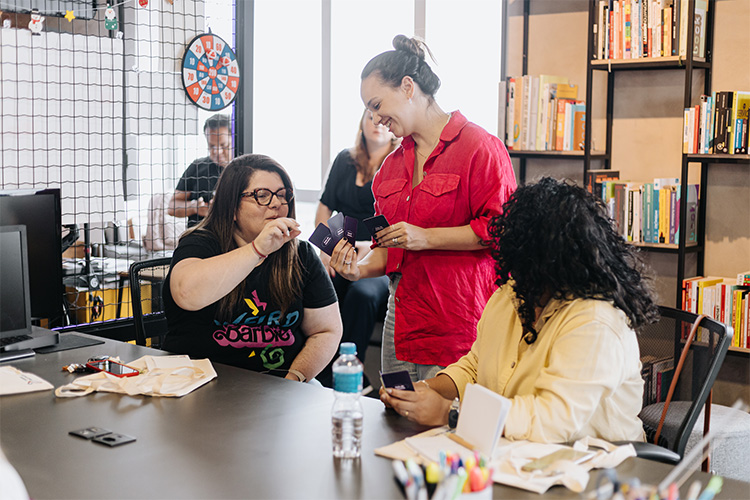 Duas mulheres conversando e trocando cartões de atividades enquanto uma terceira observa.