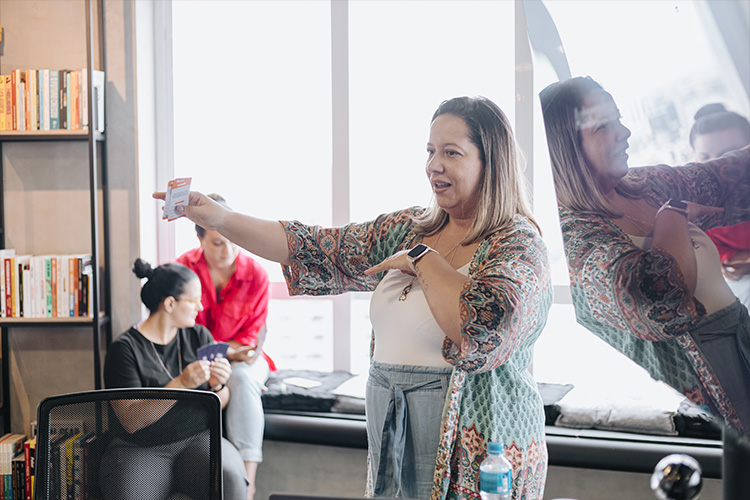 Ale Miliatti apontando para as pessoas com um cartão de atividade em uma das mãos. Seu reflexo aparece na televisão que exibe os slides de aula e ao fundo é possível ver a claridade em uma das janelas enquanto duas mulheres conversam.